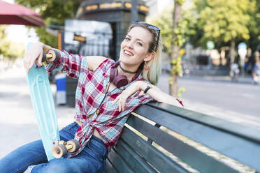 Porträt einer lächelnden jungen Frau, die auf einer Bank mit einem Skateboard sitzt - WPEF02206