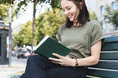 Porträt einer lächelnden jungen Frau, die auf einer Bank sitzt und ein Buch liest - WPEF02207