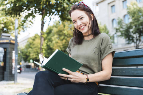 Porträt einer lächelnden jungen Frau, die mit einem Buch auf einer Bank sitzt, lizenzfreies Stockfoto
