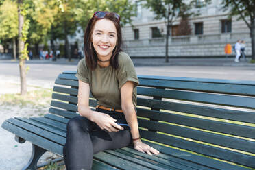 Porträt einer lächelnden jungen Frau, die mit ihrem Smartphone auf einer Bank sitzt - WPEF02209