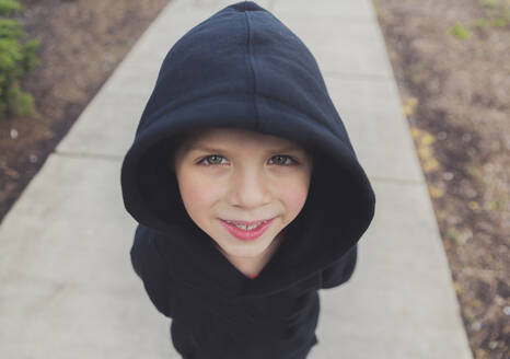 High angle portrait of happy boy wearing black hooded jacket while standing on footpath - CAVF66663