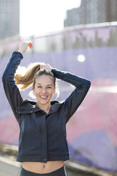 Portrait of confident athlete tying hair while standing by fence during sunny day - CAVF66711