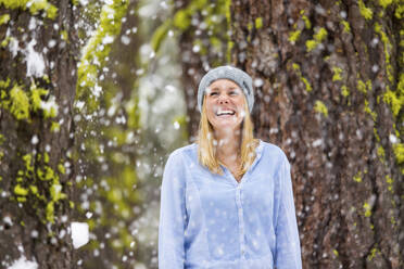 Glückliche Frau steht gegen Baumstamm bei Schneefall - CAVF66741