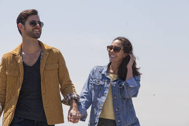 Happy couple holding hands while standing against clear sky on sunny day - CAVF66768