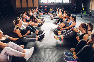 Große Gruppe von Frauen, die in einem Fitnessstudio trainieren, in Reihen auf dem Boden sitzend, mit angehobenen Beinen - CUF52925