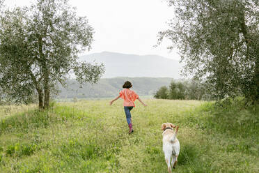 Mädchen läuft mit Labradorhund in malerischer Feldlandschaft, Rückansicht, Citta della Pieve, Umbrien, Italien - CUF52976