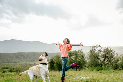 Mädchen mit offenen Armen spielt mit Labradorhund in malerischer Feldlandschaft, Citta della Pieve, Umbrien, Italien, lizenzfreies Stockfoto