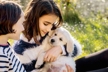 Two girls sitting in orchard hugging a cute golden retriever puppy, Scandicci, Tuscany, Italy - CUF52985