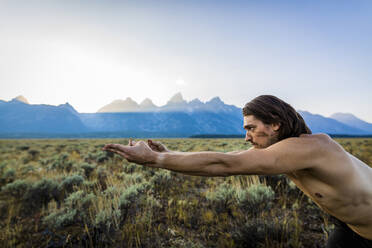 Mann ohne Hemd mit erhobenen Armen übt Yoga gegen Berge und Himmel im Bridger-Teton National Forest - CAVF66782