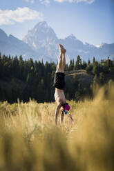 Mann ohne Hemd macht Handstand, während er auf einem Feld im Bridger-Teton National Forest Yoga gegen die Berge übt - CAVF66789