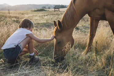 Seitenansicht eines Mädchens, das ein Pferd berührt, während es auf einer Wiese hockt - CAVF66815