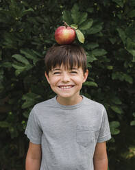 Glücklicher Junge, der draußen an einem Baum einen Apfel auf dem Kopf balanciert. - CAVF66942