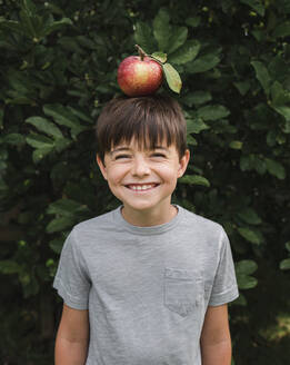 Happy young boy balancing an apple on his head outside by a tree. - CAVF66942