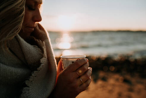 Frau hält eine Tasse Kaffee am Strand bei Sonnenuntergang - CAVF67013