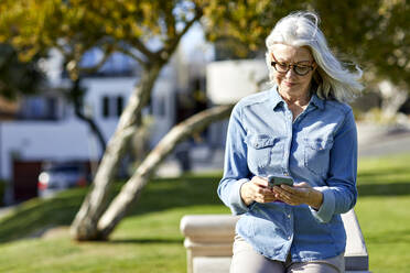 Lächelnde Frau, die ihr Smartphone benutzt, während sie an einer Stützmauer im Park steht, an einem sonnigen Tag - CAVF67113