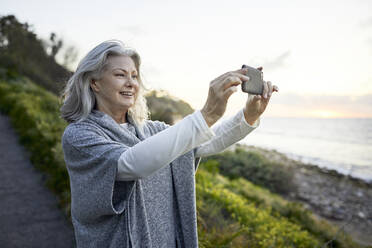 Seitenansicht einer fröhlichen älteren Frau, die mit ihrem Smartphone am Manhattan Beach bei Sonnenuntergang fotografiert - CAVF67127