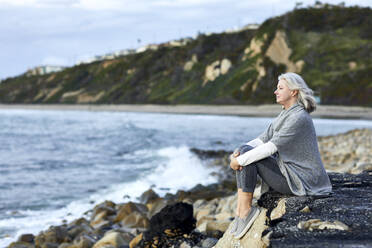 Seitenansicht einer älteren Frau, die bei Sonnenuntergang auf einem Felsen sitzend aufs Meer blickt - CAVF67129