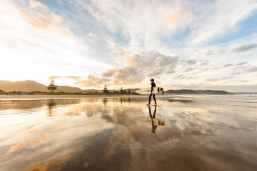 Teenager walking on beach holding surfboard at dusk - CAVF67136