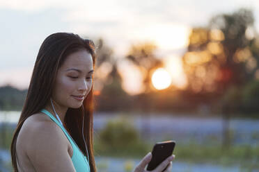 Frau, die ihr Smartphone benutzt, während sie bei Sonnenuntergang Musik hört - CAVF67182