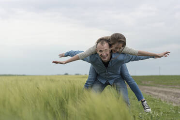 Porträt eines fröhlichen Paares mit ausgestreckten Armen, das auf einer Wiese gegen den Himmel Huckepack fährt - CAVF67408