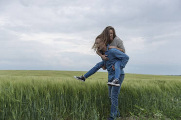 Junger Mann nimmt Frau auf grasbewachsenem Feld gegen bewölkten Himmel huckepack - CAVF67409