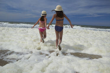 Rückansicht von Schwestern, die am Strand in Meeresschaum laufen und sich an den Händen halten - CAVF67460