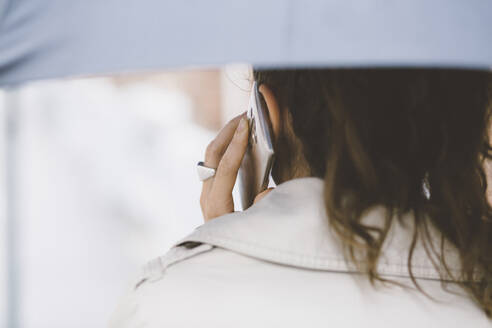 Rückansicht einer Frau mit Regenschirm, die mit einem Mobiltelefon spricht - CAVF67670