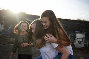 Happy teenage girl looking at friends piggybacking during sunny day - CAVF67721