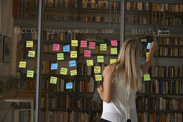 Rear view of businesswoman sticking notes on glass wall in office - CAVF67730