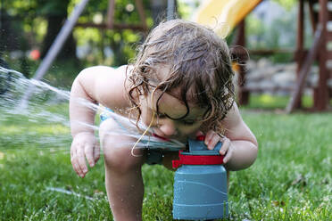 Verspieltes Mädchen trinkt Wasser aus dem Rasensprenger im Garten - CAVF67740