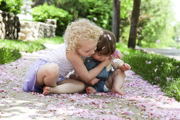 Mädchen umarmt Schwester, während sie auf dem Fußweg im Park sitzt - CAVF67756