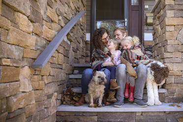 Happy family wrapped in blanket with dogs sitting on steps against house - CAVF67761