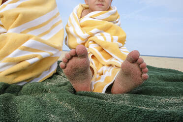 Low angle view of boy wrapped in towel while sitting at beach against sky - CAVF67781