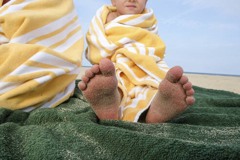 Niedriger Blickwinkel auf einen Jungen, der in ein Handtuch eingewickelt am Strand sitzt, gegen den Himmel - CAVF67781