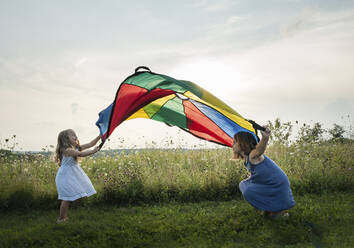 Playful Schwestern spielen mit bunten Textil auf grasbewachsenen Feld gegen den Himmel - CAVF67855