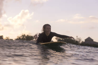 Happy woman lying on surfboard while swimming on sea against sky during sunset - CAVF67893