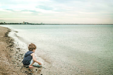 Boy holding bowl with pebbles while crouching at beach - CAVF68074