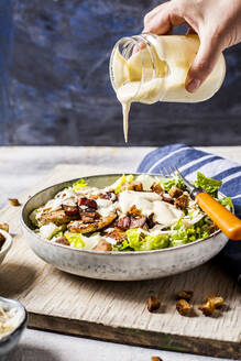 Hand of person pouring dressing over bowl of Caesar salad with romaine lettuce, Parmesan cheese, bacon, chicken breast and croutons - SBDF04080