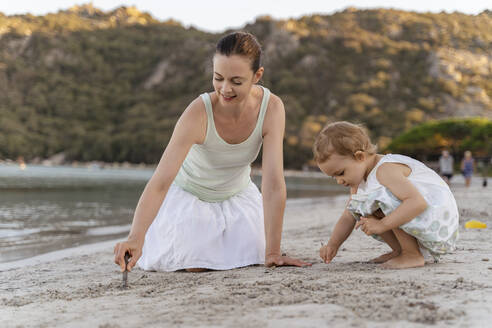 Mother and daughter drawing with little sticks in sand on the beach - DIGF08750