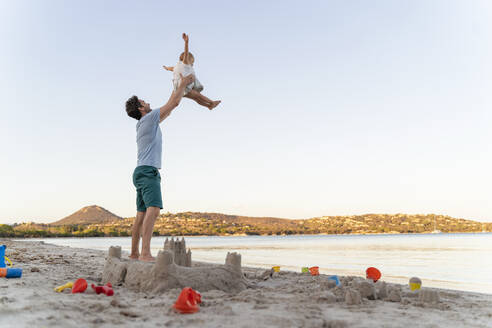 Sandburg und glücklicher Vater, der seine Tochter am Strand hochhebt - DIGF08753