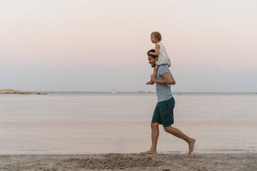 Happy father carrying daughter on shoulders on the beach at sunset - DIGF08758