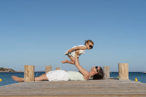 Glückliche Mutter hebt Tochter auf einem Steg am Meer hoch, lizenzfreies Stockfoto