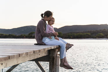 Happy mother with daughter sitting on a jetty at sunset - DIGF08778