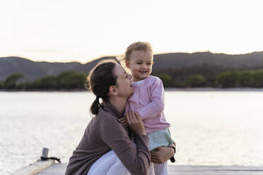 Happy mother holding her daughter on a jetty at sunset - DIGF08781