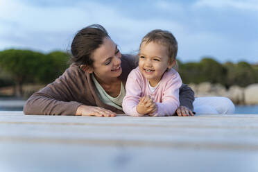 Happy mother with daughter lying on a jetty at sunset - DIGF08786