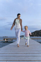 Glückliche Mutter mit Tochter, die bei Sonnenuntergang auf einem Steg spazieren gehen - DIGF08790