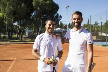 Zwei Männer in weißer Sportkleidung halten Schläger und lächeln, während sie auf einem Tennisplatz stehen - ABZF02685