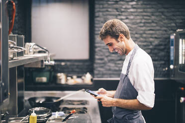 Lächelnder Koch mit Smartphone in der Küche eines Restaurants - OCMF00866