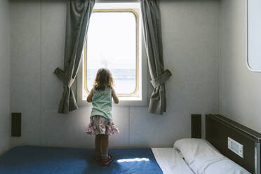 Back view of little girl standing on bed looking to the sea from window of ship's cabin - GEMF03253