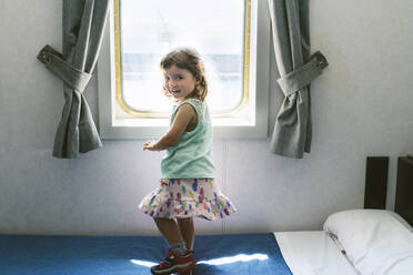 Porträt eines kleinen Mädchens, das auf dem Bett einer Schiffskabine vor einem Fenster steht - GEMF03254